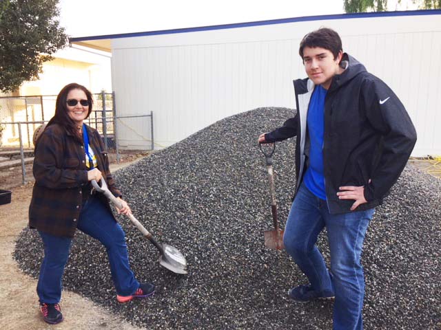 Photo of Woman digging into a pile of gravel. Standing nearby is a high school boy leaning against a shovel.